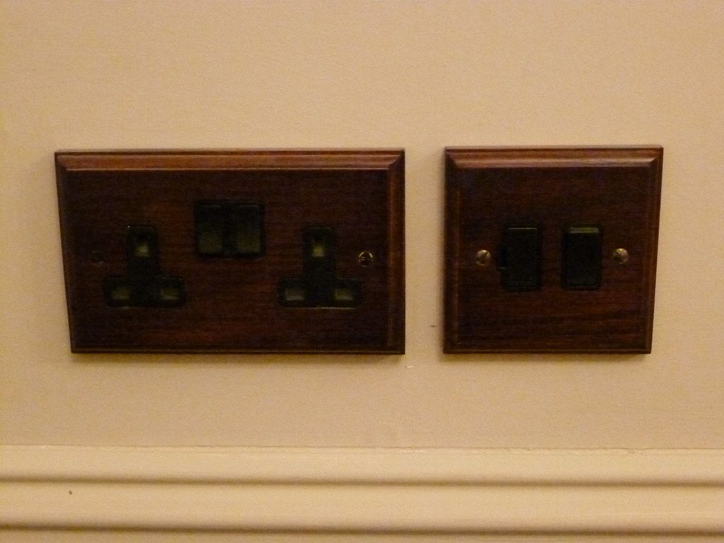A double socket and a double switch, both on dark wooden mounting plates above a painted skirting