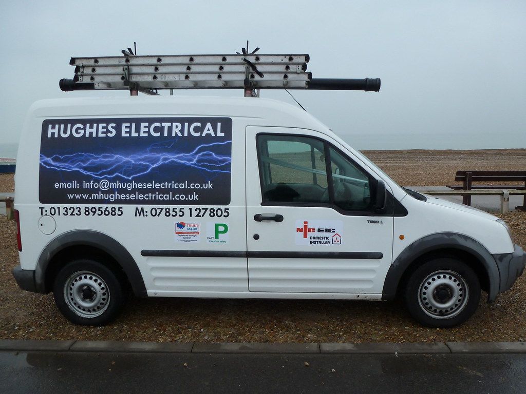 The white Hughes Electrical van with ladders on the roof rack, parked by a shingle beach, its side panel showing both phone numbers